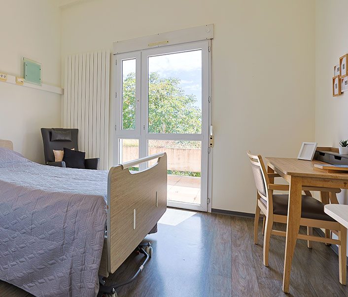 Chambre individuelle avec lit médicalisée, télévision, fauteuil et table, donnant sur le jardin